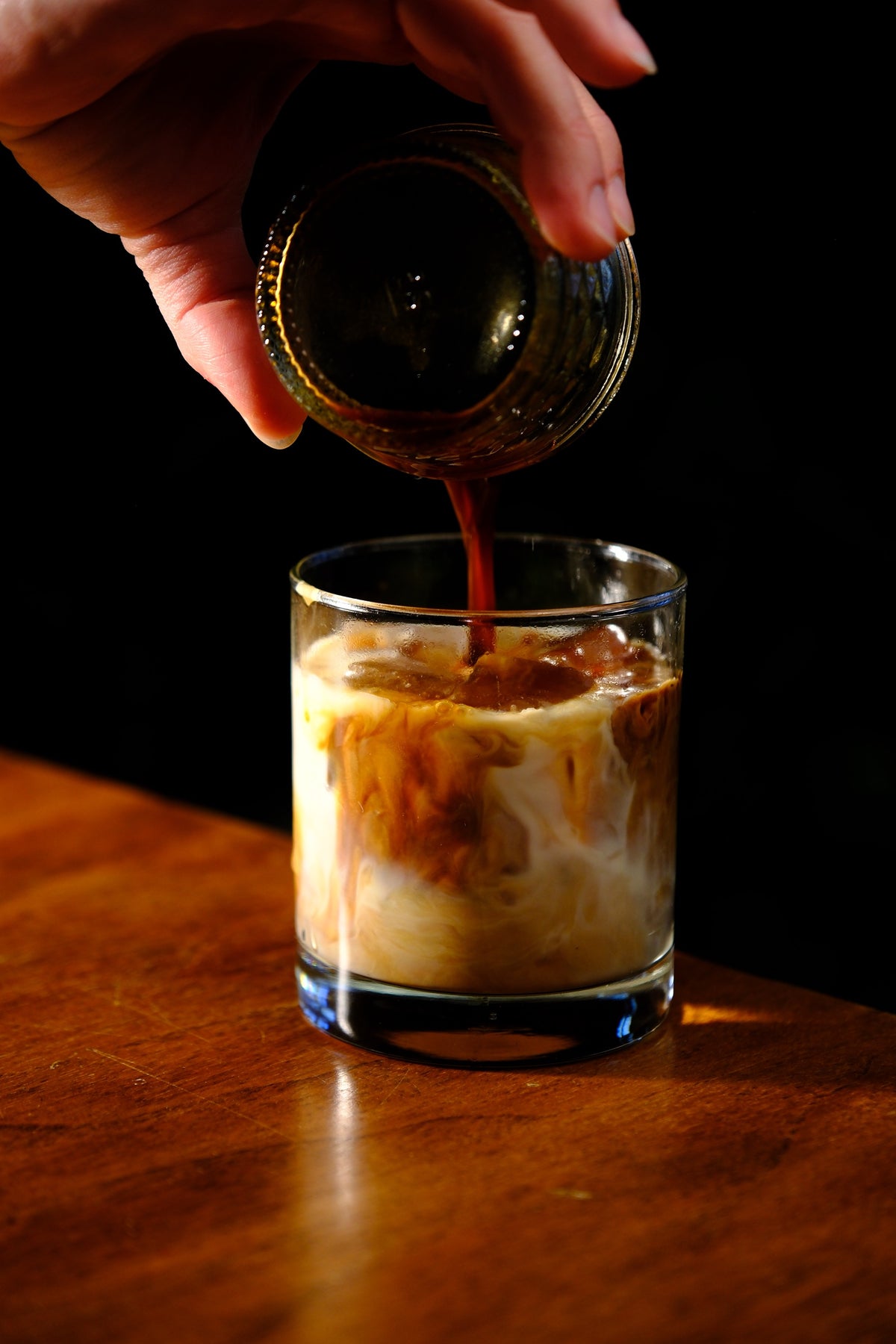 A hand pouring a shot of espresso, into a clear glass tumbler containing a creamy off-white drink, set on a wooden table against a dark background.